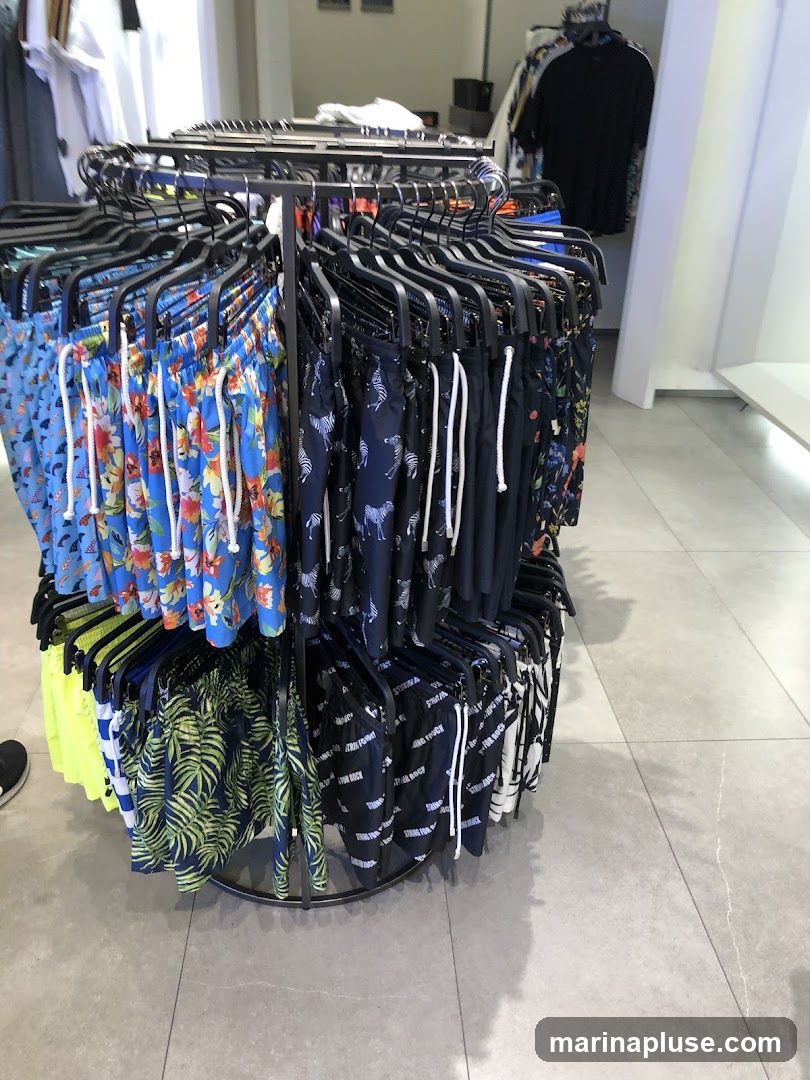 A circular rack displays a variety of men's swim trunks with different patterns, including floral, zebra, palm leaf, and text prints. The swim trunks are hung on black hangers. In the background, clothing racks with shirts and other garments are visible, along with a white wall and a light-colored floor.