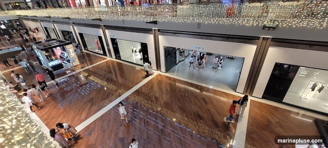 An overhead view of several Zara storefronts within a brightly lit mall. People are seen walking through the mall and inside the stores. The storefronts feature large glass windows displaying mannequins and clothing. The mall has a polished wooden floor and is decorated with strings of lights along the upper levels.