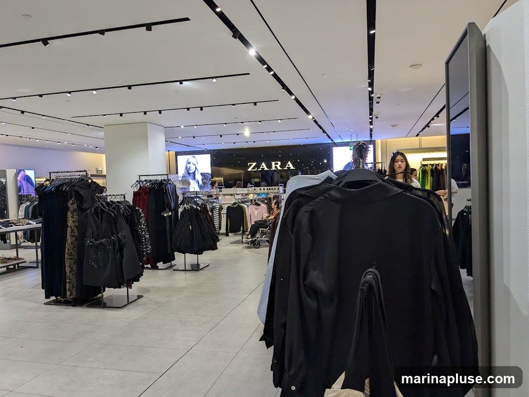 The interior of a Zara clothing store. Racks of women's clothing are displayed on the left and center of the image. A large screen displaying a model is visible in the background, along with the Zara logo. Several shoppers are browsing the merchandise.