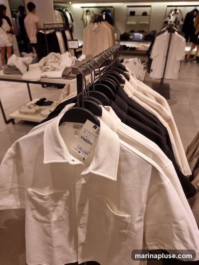 A rack of short-sleeved shirts hangs in the foreground of a clothing store. The shirts are white and black. In the background, other clothing items are displayed on racks and shelves, and shoppers can be seen browsing. A price tag on one of the white shirts shows "€29.95".