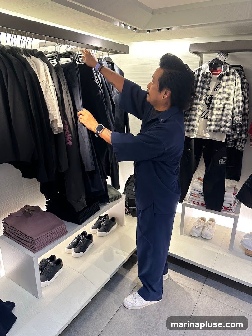 A man in a navy blue uniform is organizing clothes on a rack in a store. The rack has various items of clothing, including black pants and shirts. Below the rack, there are shelves displaying folded shirts and pairs of black sneakers. To the right, there is another display with a plaid shirt and pants, as well as stacks of t-shirts and more sneakers. The store appears to be well-lit with modern shelving and lighting fixtures.