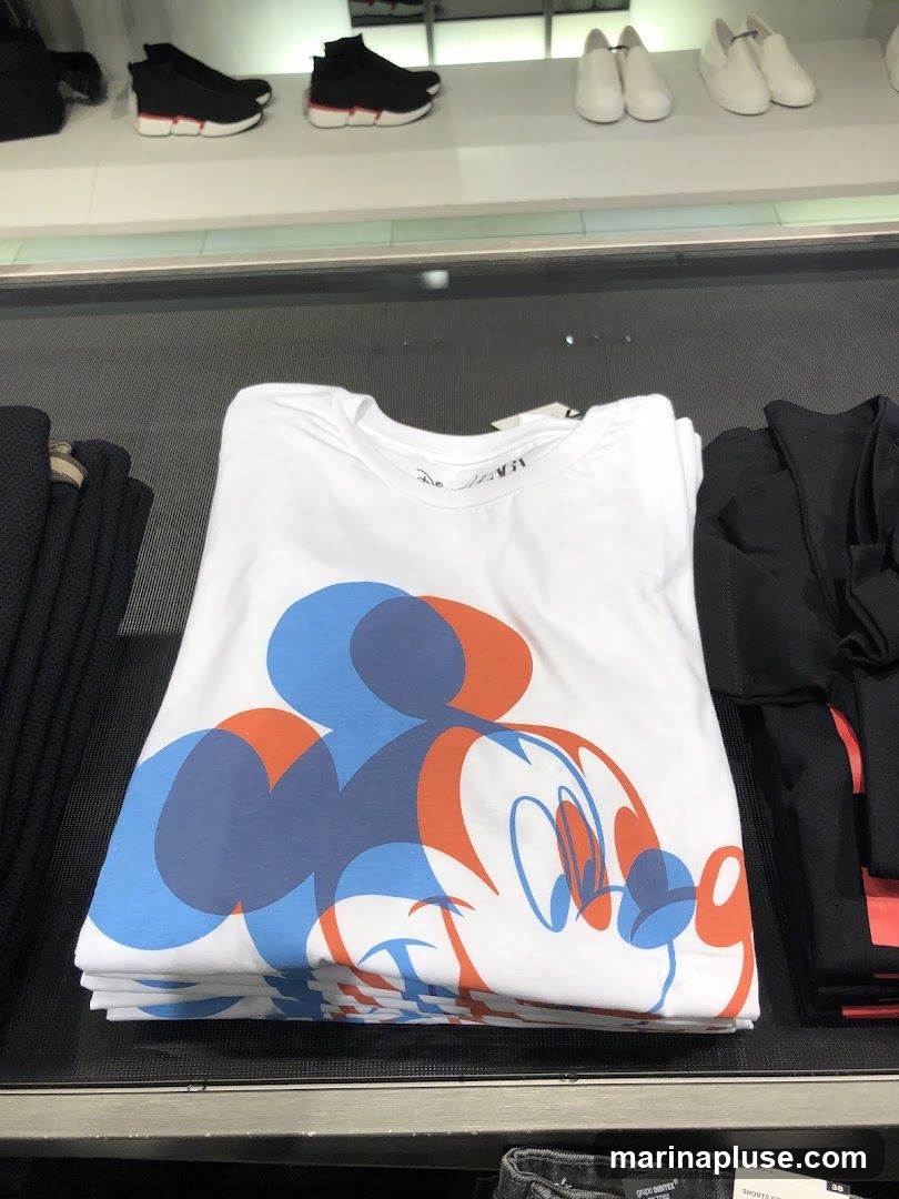 A folded white t-shirt with a large graphic of Mickey Mouse's face, rendered in blue and orange overlapping outlines. The t-shirt is displayed on a counter, with other clothing items visible on shelves above and to the sides.