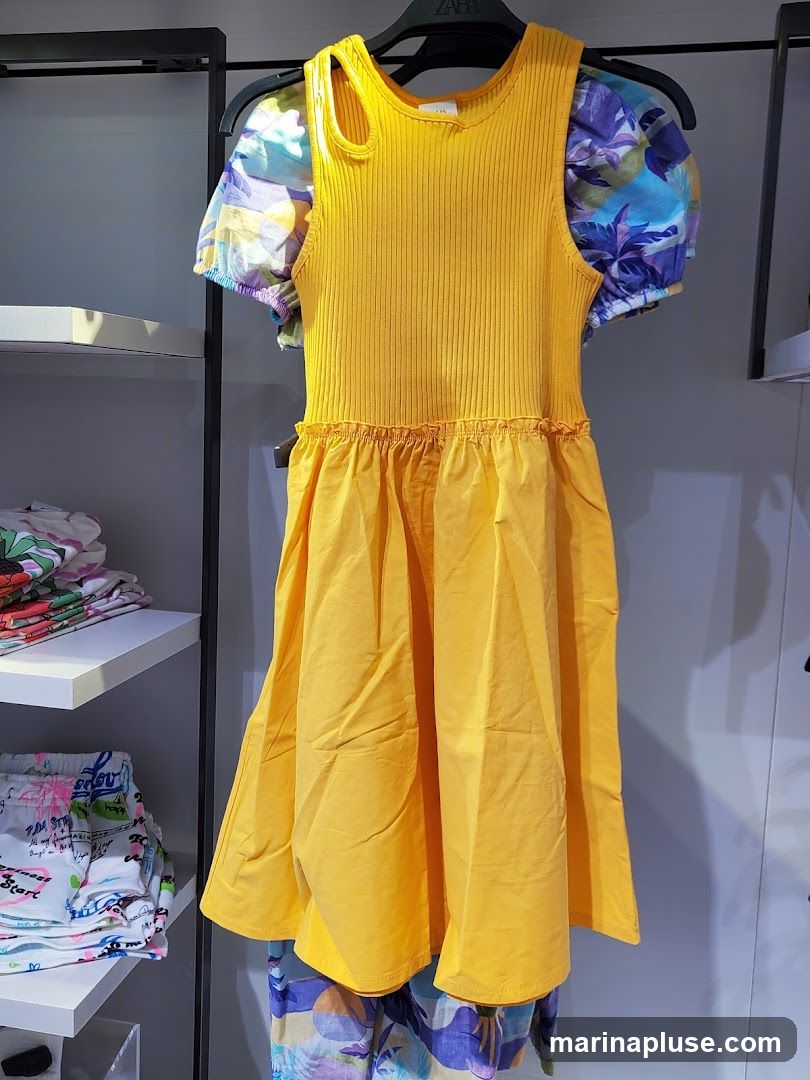 A yellow, ribbed sleeveless dress with a ruffled waist and a full skirt is displayed on a hanger. The dress has short, puffed sleeves with a colorful floral print. Below the dress, a pair of patterned pants with a similar floral design is visible. The clothing is on display in a store, with shelves of folded clothing in the background. A hanger with the brand name "ZARA" is visible.