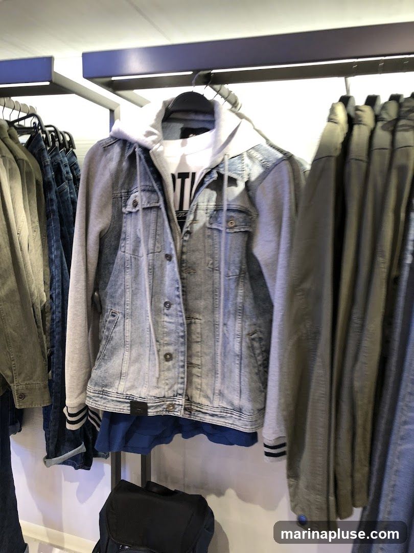 A light wash denim jacket with grey sleeves and a white t-shirt underneath is displayed on a hanger. The jacket has a hood attached and is part of a rack of clothing, including jeans and other jackets. A dark backpack is visible at the bottom of the image.