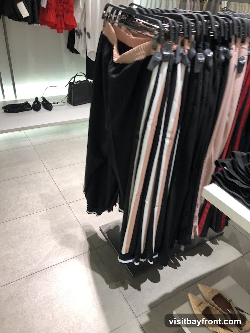 A rack of black pants with striped accents is displayed in a store. The pants have a light pink and white stripe down the side. In the background, there are other clothing items and a black handbag. On the floor, there are several pairs of shoes.