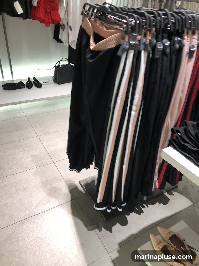 A rack of black pants with striped accents is displayed in a store. The pants have a light pink and white stripe down the side. In the background, there are other clothing items and a black handbag. On the floor, there are several pairs of shoes.