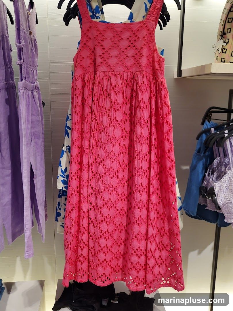 A bright pink sleeveless dress with a square neckline and broderie anglaise detailing is displayed on a hanger. The dress has a gathered waist and a full, midi-length skirt. To the left, a lavender dress is visible, and to the right, other garments are hanging on racks.