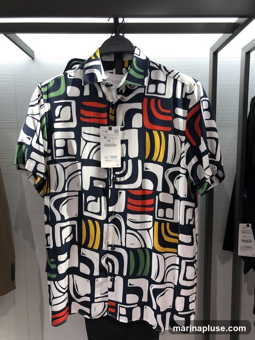 A short-sleeved collared shirt with a bold, abstract print in navy, white, red, yellow, and green. The shirt is hanging on a hanger in a retail environment. A price tag is visible, showing a price of 59.90.