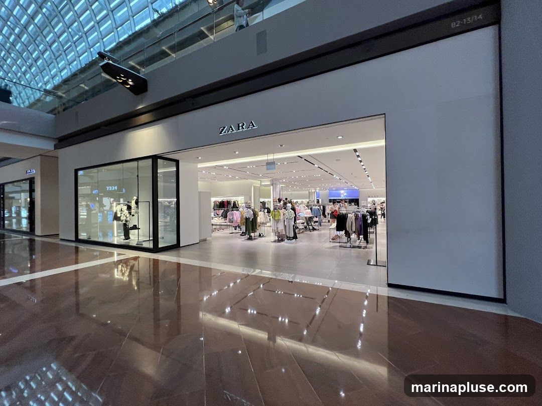 The exterior of a Zara clothing store in a mall. The store has a large glass window display on the left and an open entrance to the right. The Zara logo is visible above the entrance. Inside, racks of clothing are displayed. The floor is polished and reflects the store and ceiling lights.