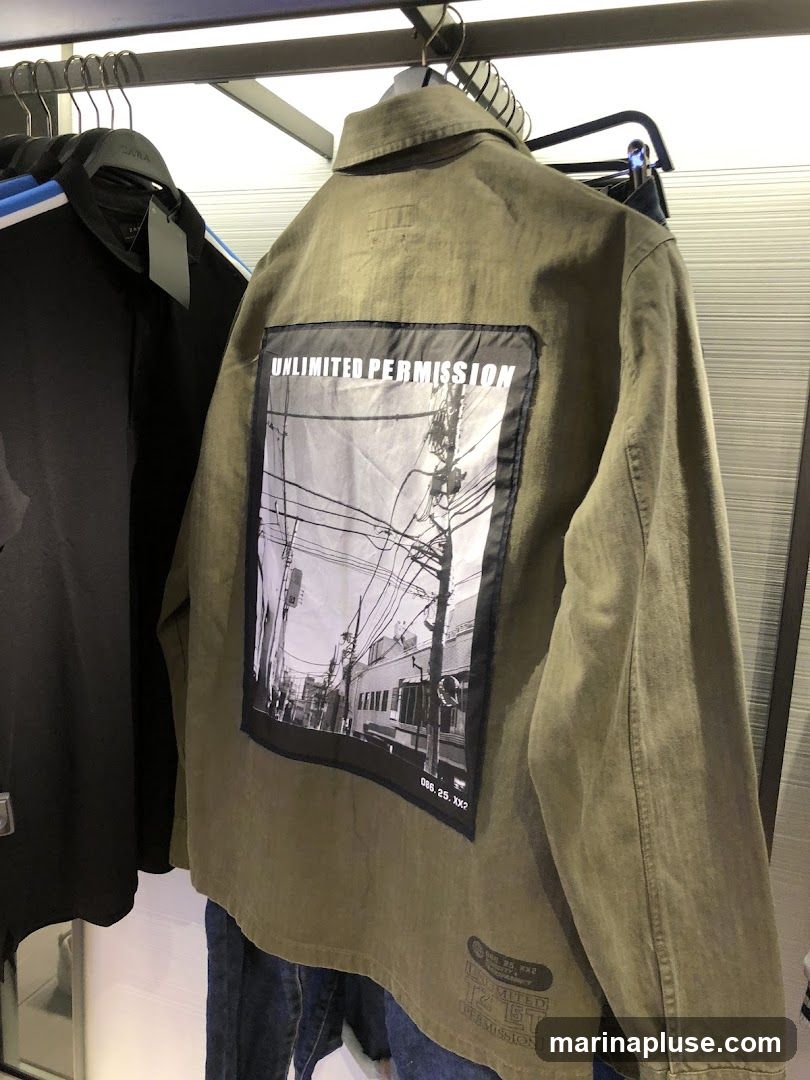 A close-up shot of the back of a green jacket with a large black and white graphic print. The print features a cityscape with power lines and buildings. Text on the print reads "UNLIMITED PERMISSION" and "086. 25. XX?". Below the graphic, there is a smaller print with text that includes "UNLIMITED PERMISSION" and "12 ST". The jacket is hanging on a rack with other clothing items, including a black jacket with blue stripes.
