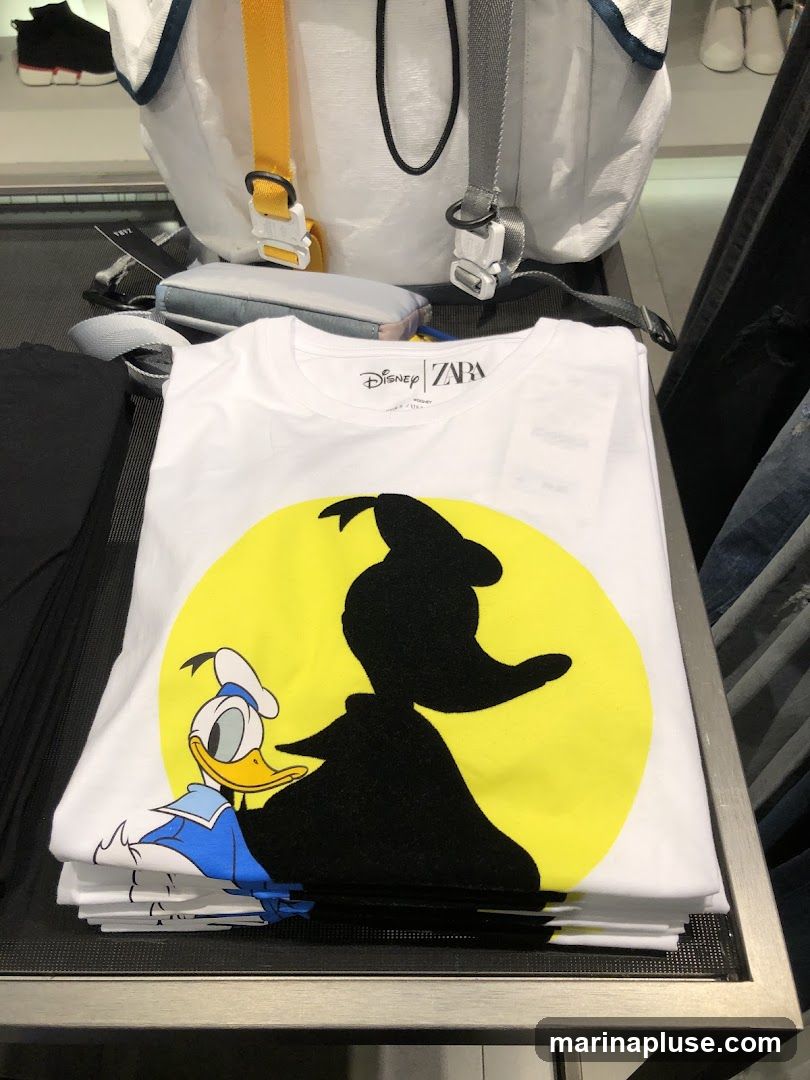 A white t-shirt with a graphic print of Donald Duck and his silhouette against a yellow circle. The t-shirt is folded and stacked with other t-shirts. The Disney and Zara logos are visible on the neckline. In the background, a white backpack with yellow and grey straps is visible, along with some clothing items on shelves.