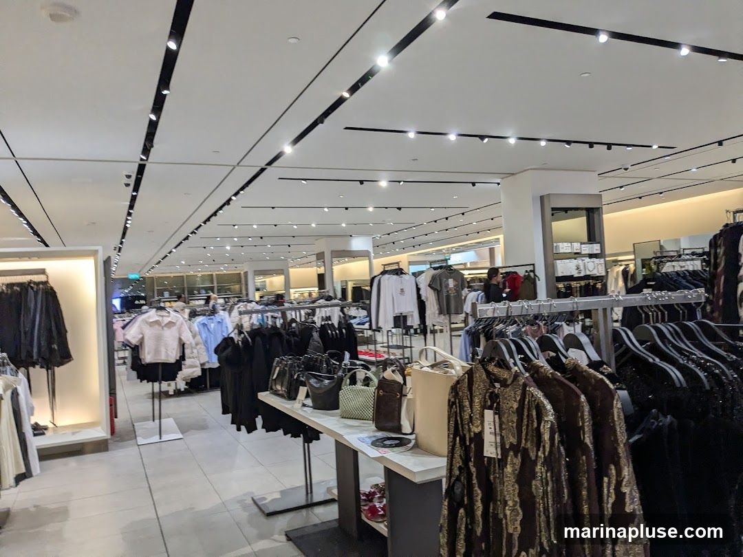 A well-lit clothing store interior with racks of apparel and accessories. The store features modern lighting and displays, showcasing a variety of clothing items including jackets, shirts, and dresses, as well as handbags.
