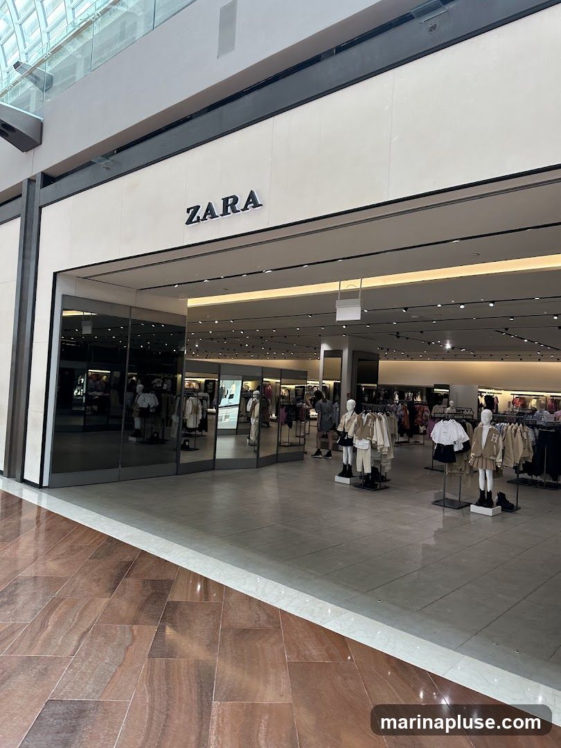 The exterior entrance of a Zara store with the brand name clearly visible above the entrance. The store interior, displaying clothing on mannequins and racks, is visible through the large glass windows.