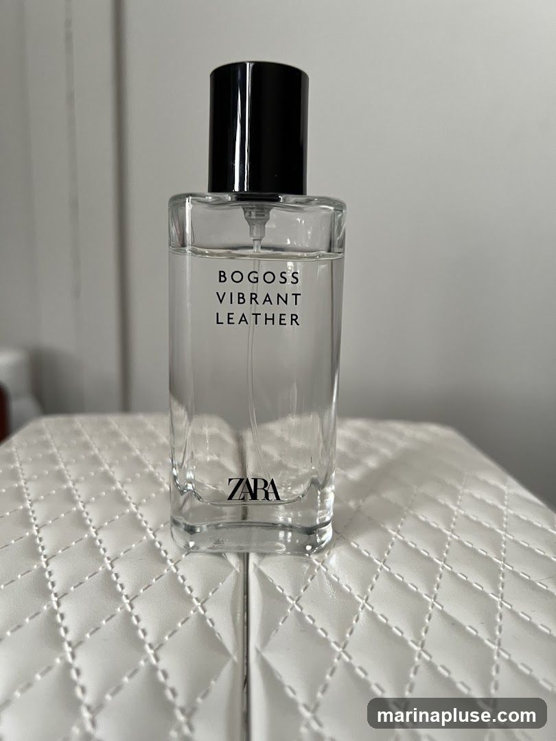 A clear glass perfume bottle with a black cap sits on a white quilted surface. The bottle has black text that reads "BOGOSS VIBRANT LEATHER" and the Zara logo at the bottom.