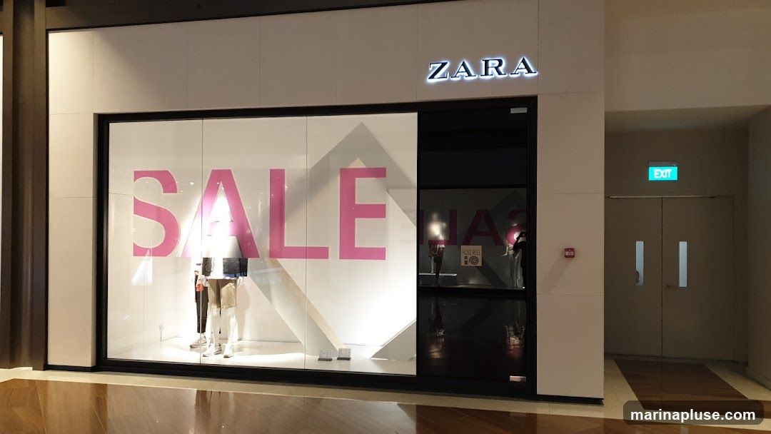 The exterior of a Zara store with a large window displaying the word "SALE" in pink lettering. Mannequins are visible inside the window. The Zara logo is illuminated above the entrance. To the right of the entrance are double doors with an illuminated green exit sign above them.