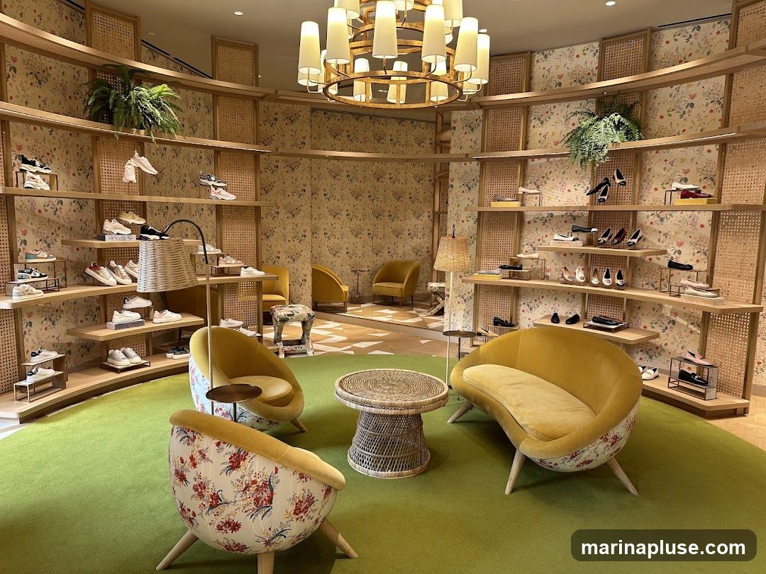 A well-lit and elegantly designed shoe store interior with a circular display of shelves showcasing various footwear. The shelves are made of light-colored wood and are set against a floral-patterned wallpaper with woven cane accents. Several pairs of sneakers and dress shoes are neatly arranged on the shelves. In the center of the room, there are comfortable seating areas with mustard-yellow velvet chairs and sofas, complemented by small woven coffee tables. A large, ornate chandelier hangs from the ceiling, adding to the luxurious ambiance. The floor is covered with a vibrant green carpet.