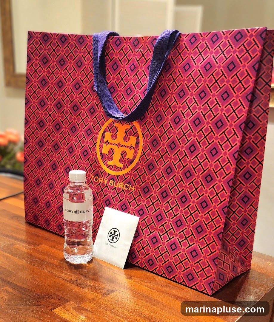 A large Tory Burch shopping bag with a repeating geometric pattern in pink and purple. The Tory Burch logo is prominently displayed in gold on the front of the bag. Next to the bag are a small bottle of water with a Tory Burch label and a small white packet also bearing the Tory Burch logo. The items are placed on a wooden surface.