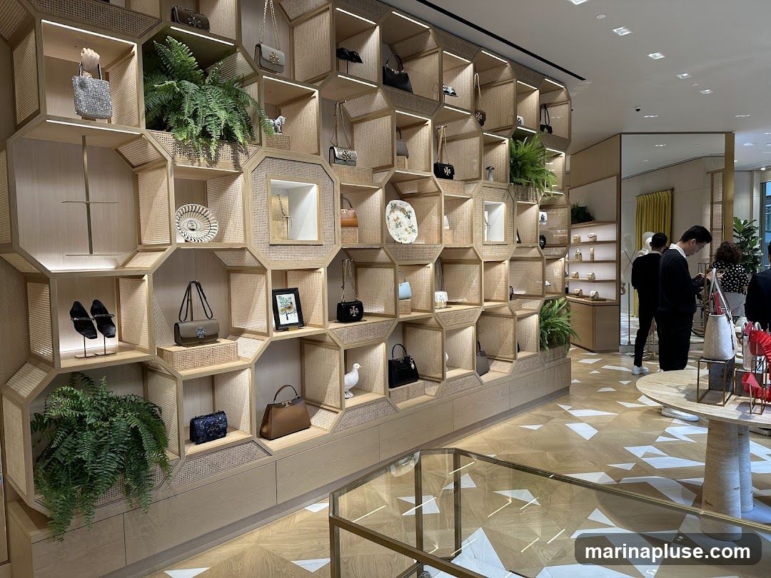 The interior of a store features a large, hexagonal display shelving unit filled with handbags, shoes, decorative plates, and plants. The shelves are made of light-colored wood and woven cane. In the background, several customers are browsing, and there are additional display shelves and tables. The floor has a geometric wood pattern.