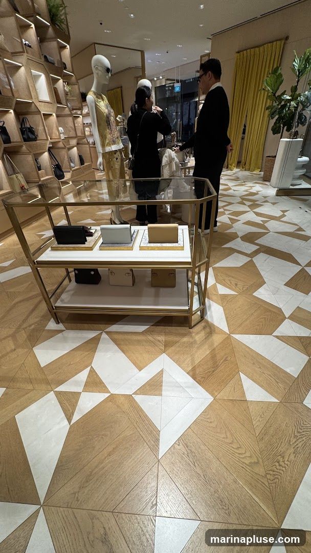 The image shows the interior of a store with a mannequin displaying a patterned top and sunglasses. Behind the mannequin, shelves are filled with handbags. A display case in the foreground holds several clutch bags. Two people are visible in the background, one appearing to be a sales associate assisting a customer. The floor has a geometric pattern of wood and white tiles. Yellow curtains hang on the right side of the image, and a potted plant is also visible.