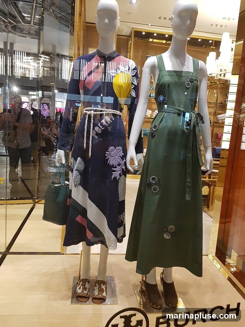 Two mannequins are displayed in a store window. The mannequin on the left wears a long-sleeved dress with a geometric and floral print, a rope belt, and brown and white studded sandals. It also holds a dark green handbag. The mannequin on the right wears a sleeveless green dress with floral embellishments and dark brown clogs. The Tory Burch logo is visible at the bottom of the display.