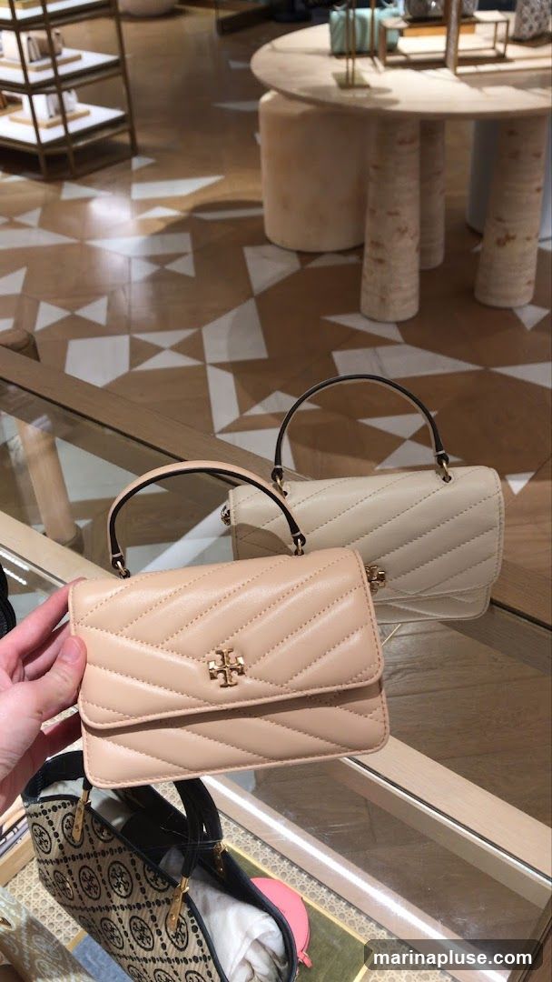 A close-up shot of two quilted handbags with top handles and gold-tone hardware, displayed on a glass counter in a retail store. The handbag in the foreground is a light beige color, and the one behind it is a cream color. Both feature the brand's signature logo. Below, another patterned tote bag is visible. The store interior features a geometric tiled floor and modern display tables.