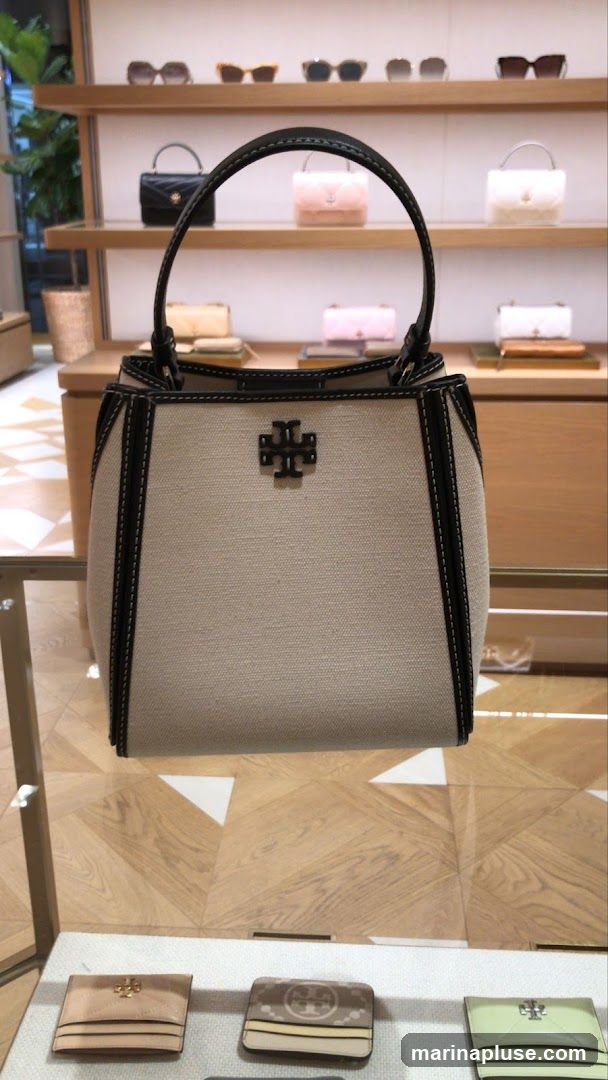 A close-up view of a light-colored canvas handbag with black trim and stitching, featuring the Tory Burch logo in the center. The handbag is displayed in a retail setting, with shelves in the background holding various other handbags and sunglasses. Below the handbag, several small accessories, possibly wallets or cardholders, are arranged on a display shelf. The flooring is a light wood pattern.