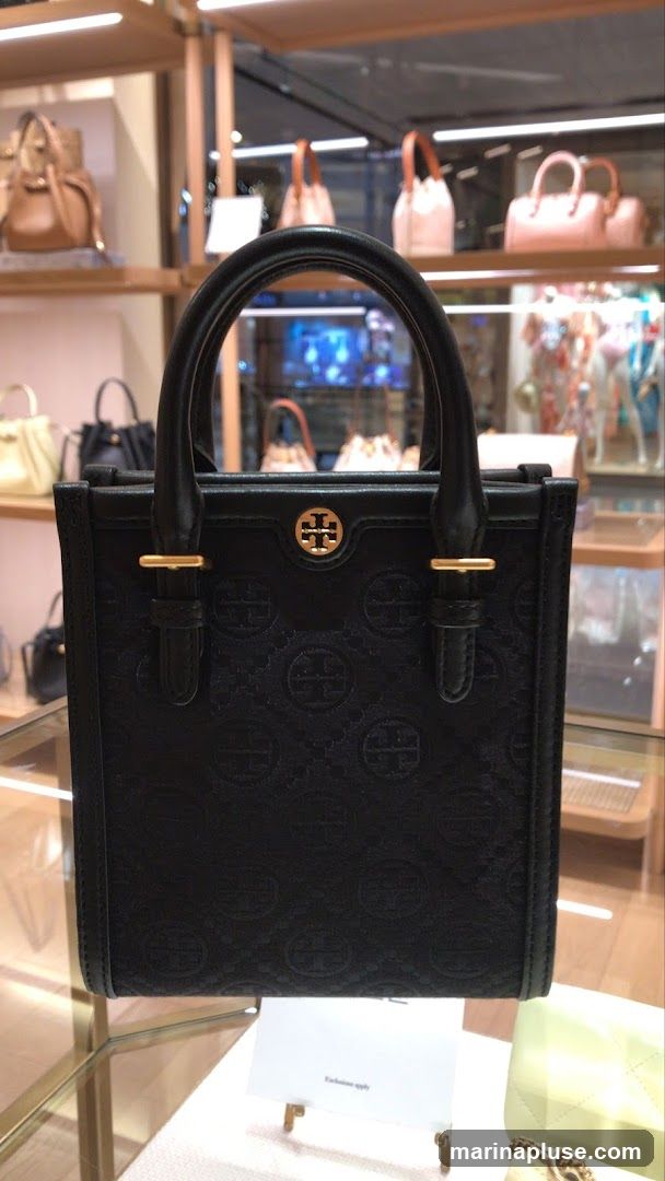A black tote bag with an embossed pattern of the brand's logo is displayed in a store. The bag has double top handles and gold-tone hardware. In the background, other handbags are visible on shelves and in display cases. A small white card with the text "Exclusive apply" is placed in front of the bag.