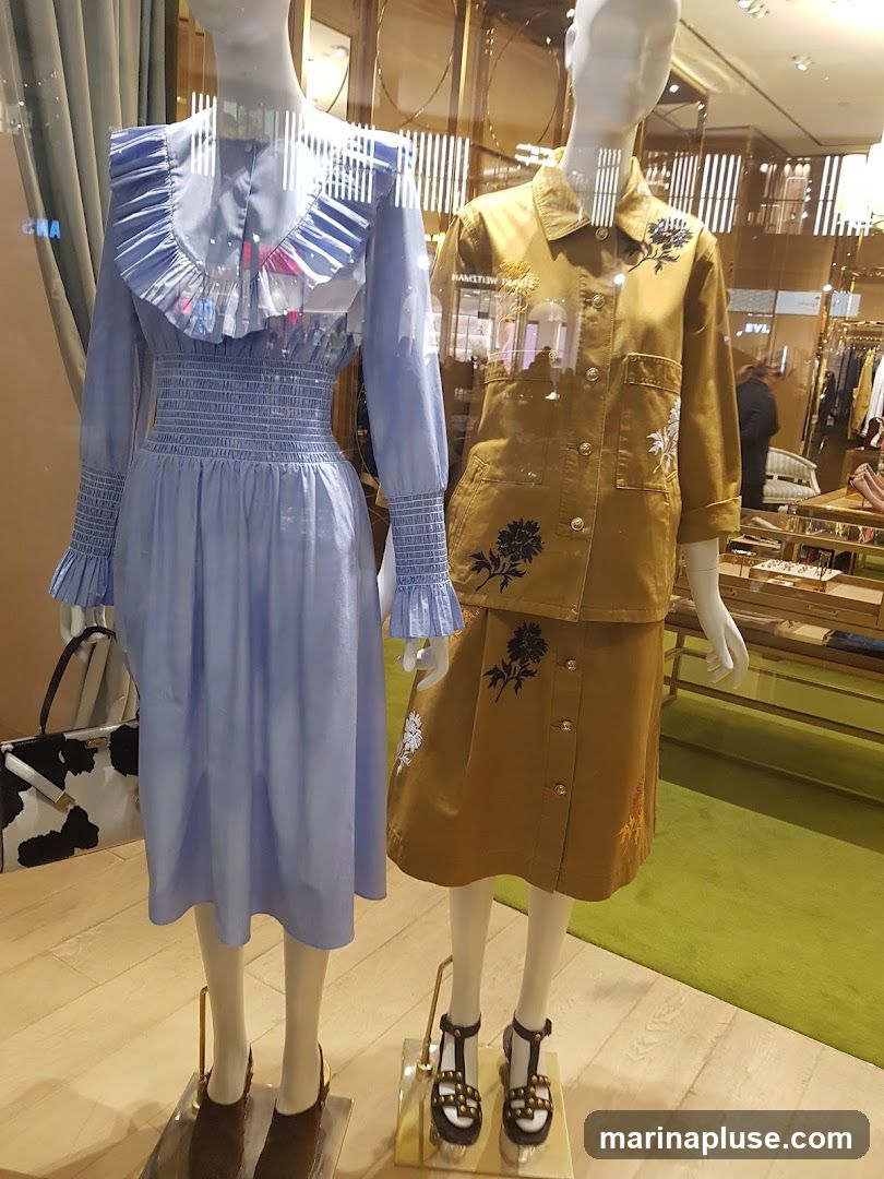 Two mannequins are displayed in a store window. The mannequin on the left wears a light blue dress with a ruffled collar and smocked waist. A black and white cow-print handbag is placed next to it. The mannequin on the right wears a tan jacket and skirt set, both adorned with floral embroidery. The background shows the interior of a store with clothing racks and displays.