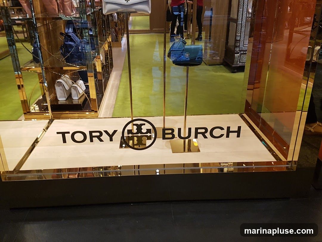 A store window display for Tory Burch featuring handbags on stands. The brand name "TORY BURCH" is prominently displayed on the floor of the window. The display includes mirrored surfaces and a green carpeted floor.