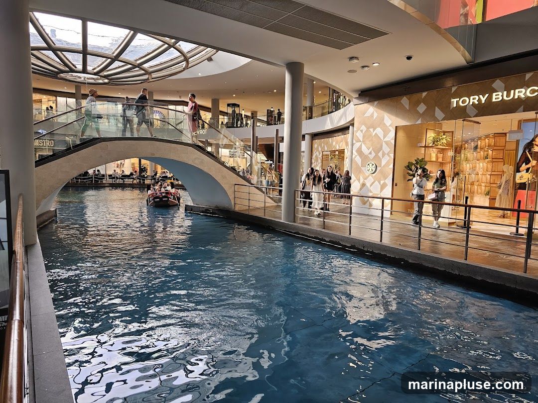 An indoor canal with clear blue water, reflecting the surrounding architecture. A gondola boat with passengers navigates the canal. People are seen walking on a bridge and along a walkway. In the background, various shops are visible, including one with the prominent signage 'TORY BURCH'. The overall scene depicts a shopping mall with a unique water feature.