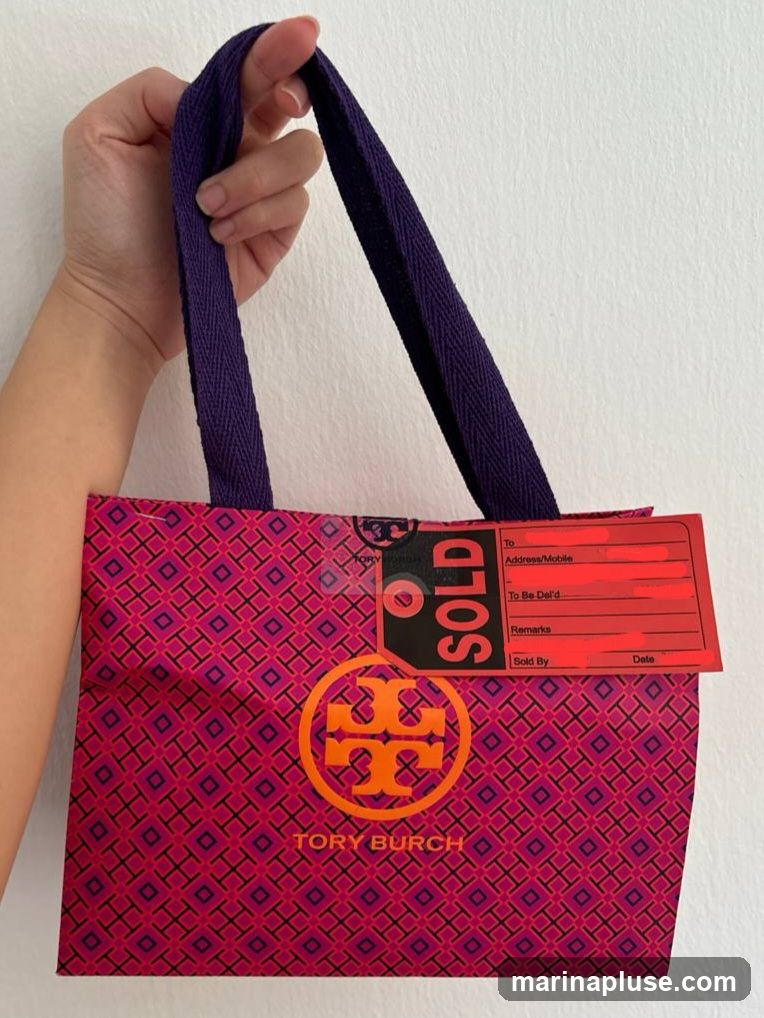 A hand holds a pink and purple patterned Tory Burch shopping bag. The bag features the Tory Burch logo and name. A red tag with the word "SOLD" is attached to the bag.