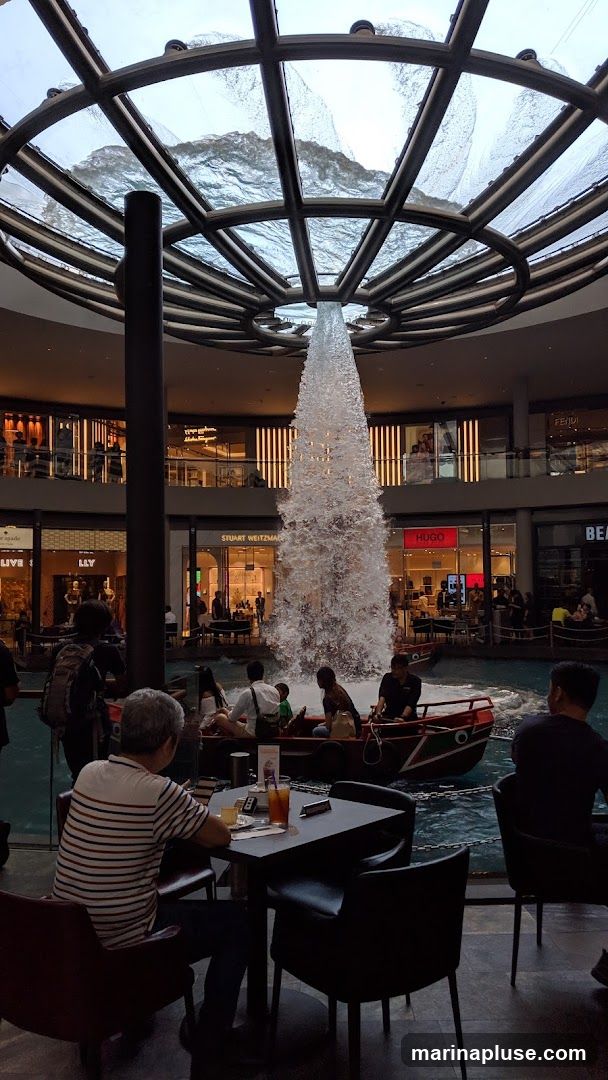 An indoor scene featuring a large, cascading waterfall in the center, with a boat carrying passengers navigating the water below. Several people are seated at tables in the foreground, appearing to be dining or relaxing. The background shows multiple levels of shops with visible signage, including Stuart Weitzman and Hugo. The architecture includes a circular, glass-paneled ceiling with metal framing, through which water is flowing.