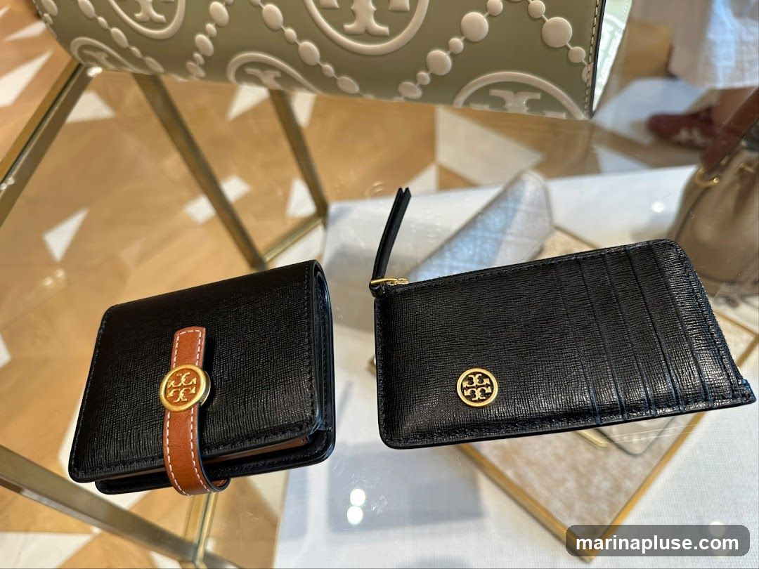 Two black Tory Burch wallets are displayed on a glass surface. The wallet on the left is a bifold wallet with a brown strap and gold logo. The wallet on the right is a cardholder with a zipper and gold logo. A light green bag with the Tory Burch logo is visible in the background.