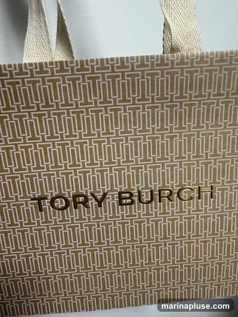 A close-up shot of a Tory Burch shopping bag. The bag is gold with a repeating pattern of white 'T' logos. The words 'TORY BURCH' are printed in gold foil in the center of the bag. The bag has beige fabric handles.