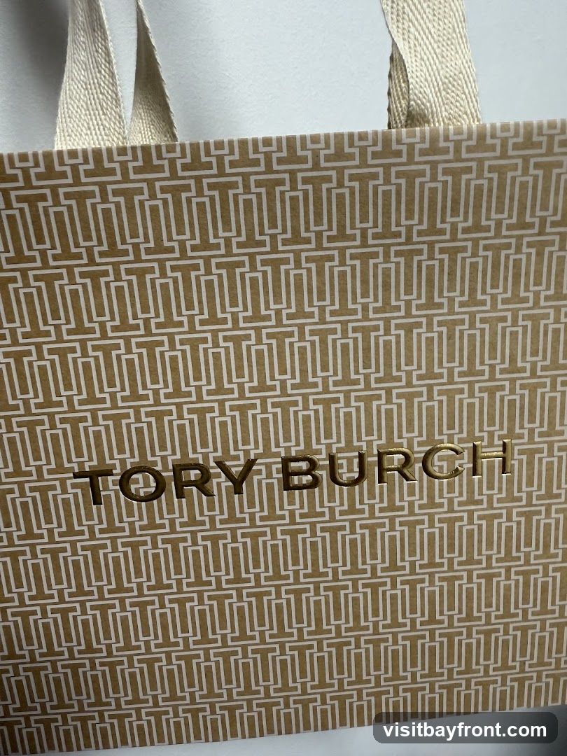 A close-up shot of a Tory Burch shopping bag. The bag is gold with a repeating pattern of white 'T' logos. The words 'TORY BURCH' are printed in gold foil in the center of the bag. The bag has beige fabric handles.