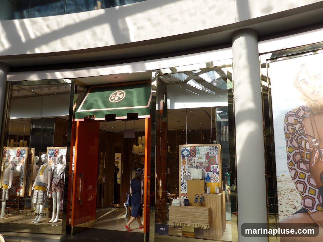 The exterior of a Tory Burch store with mannequins displaying clothing in the window. An open orange door leads into the store, and a woman in a blue dress walks inside. A large advertisement featuring a woman is on the right side of the storefront.