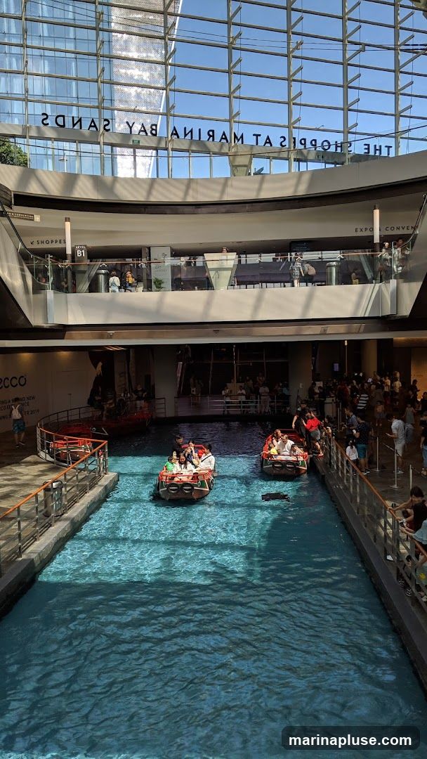People are riding in small boats on a canal that runs through a large, modern shopping mall. The mall has multiple levels with shops and escalators visible. The ceiling is a glass structure, allowing natural light to enter. The text "THE SHOPPES AT MARINA BAY SANDS" is visible reflected on the glass ceiling.