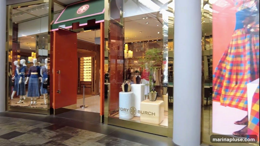 The exterior of a Tory Burch store with mannequins dressed in blue and white plaid outfits displayed in the window. The store features a large orange door and a green awning with the Tory Burch logo. Inside, clothing racks and handbags are visible. A large advertisement featuring a person in a colorful plaid skirt is on the right side of the storefront.