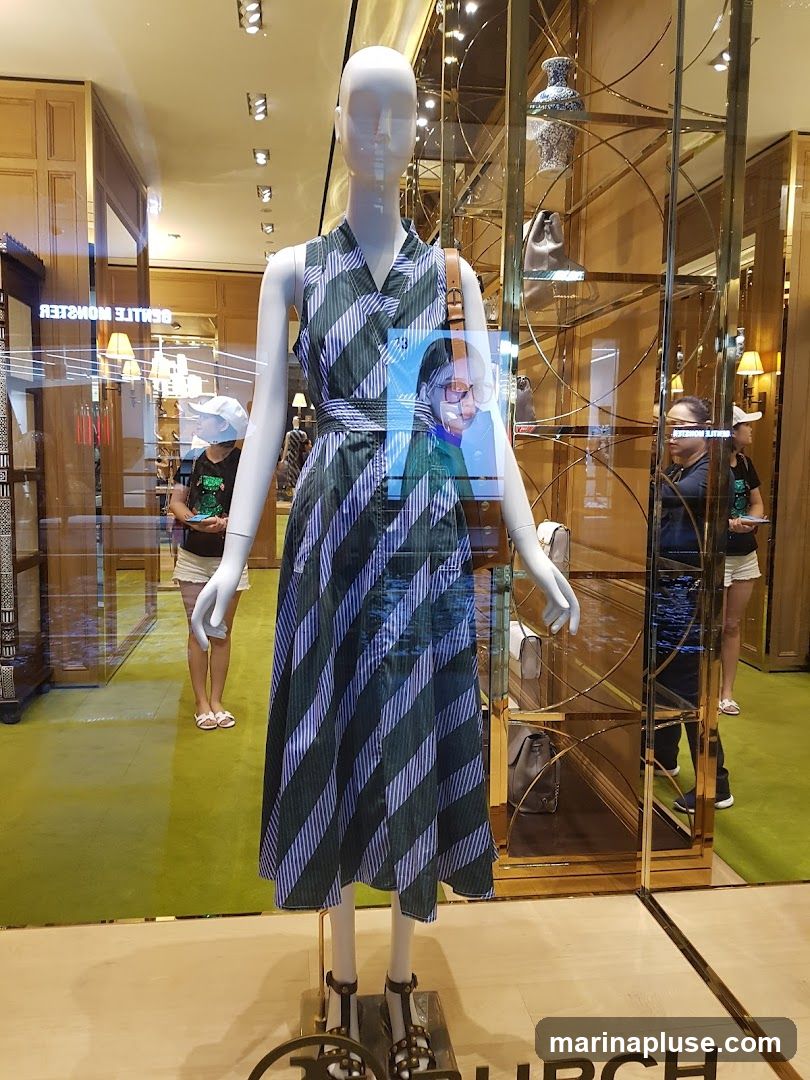 A mannequin in a striped dress is displayed in a store window. The mannequin is wearing a green and blue striped dress with a belt and brown shoulder bag. It is also wearing black sandals. In the background, there are shelves with handbags and decorative vases. Two women are visible in the reflection of the window, one wearing a white hat and the other a black shirt. The floor is covered in a green carpet. The word "BURCH" is visible on the floor in front of the display.