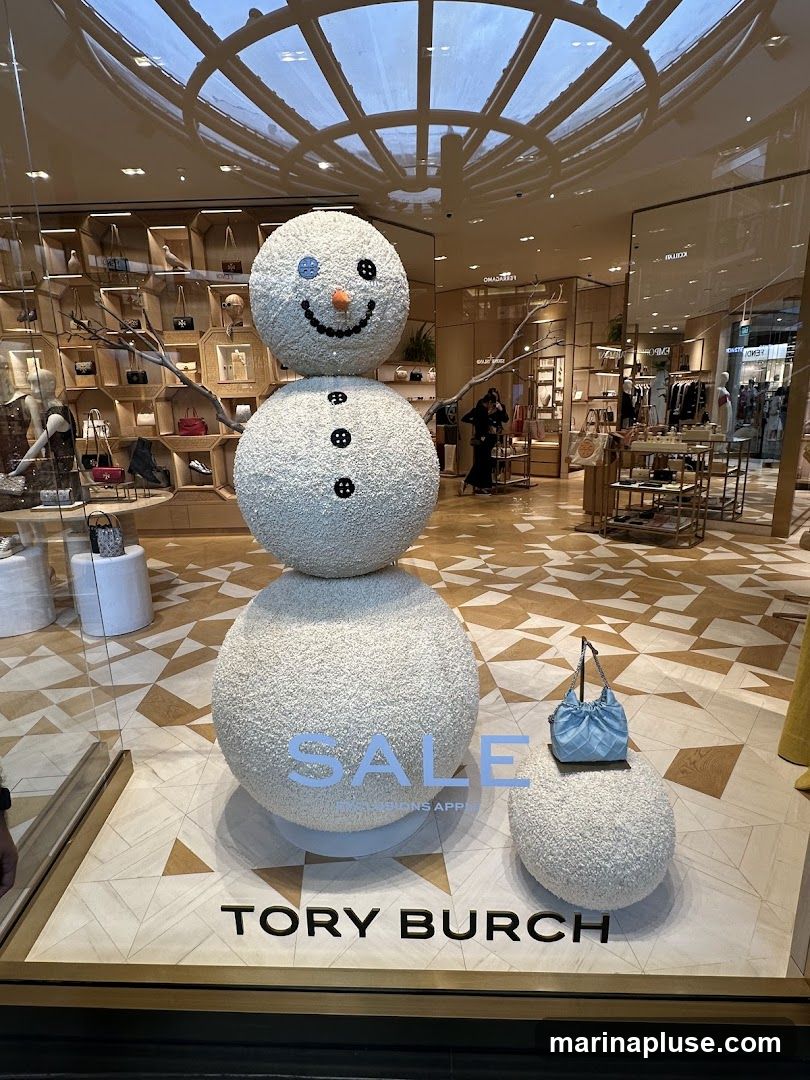 A festive store window display features a large, fluffy snowman decoration. The snowman has a smiling face with a blue button eye and an orange carrot nose. It is adorned with black buttons down its body. The word "SALE" is prominently displayed on the snowman's base, with smaller text below indicating "CONDITIONS APPLY." A small blue handbag is placed on a fluffy stool next to the snowman. The store's name, "TORY BURCH," is visible on the floor of the display. The background shows the interior of the store, with shelves displaying handbags and other merchandise.