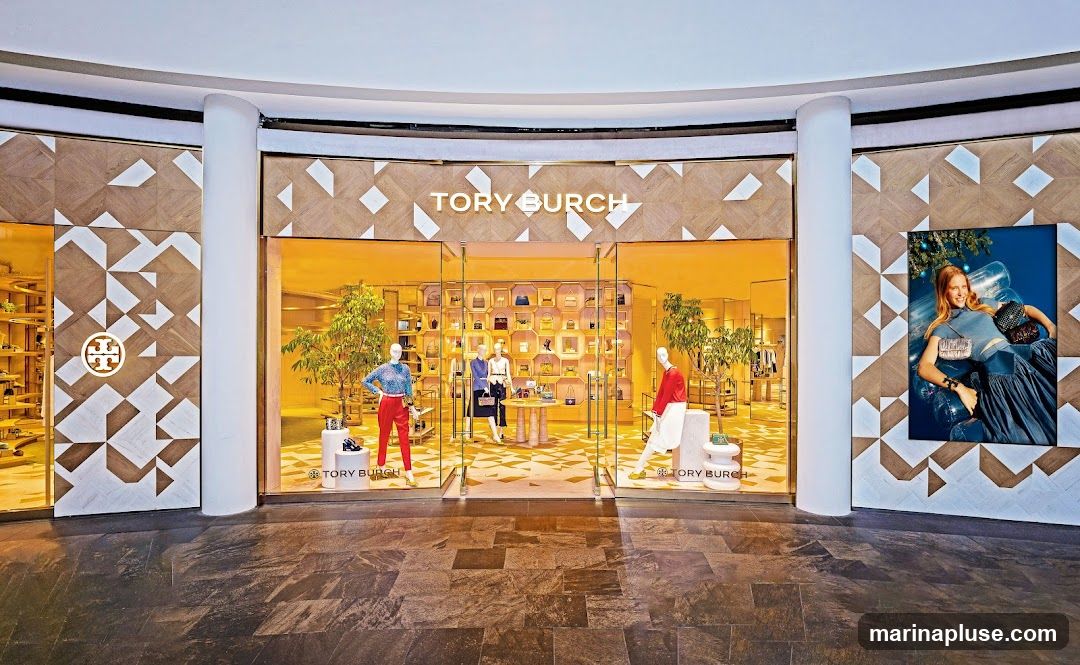 The exterior of a Tory Burch store with a large glass entrance. The store's name is prominently displayed above the entrance. Inside, mannequins are dressed in the brand's clothing, and shelves are filled with handbags. A large advertisement featuring a woman is displayed on the right side of the storefront.