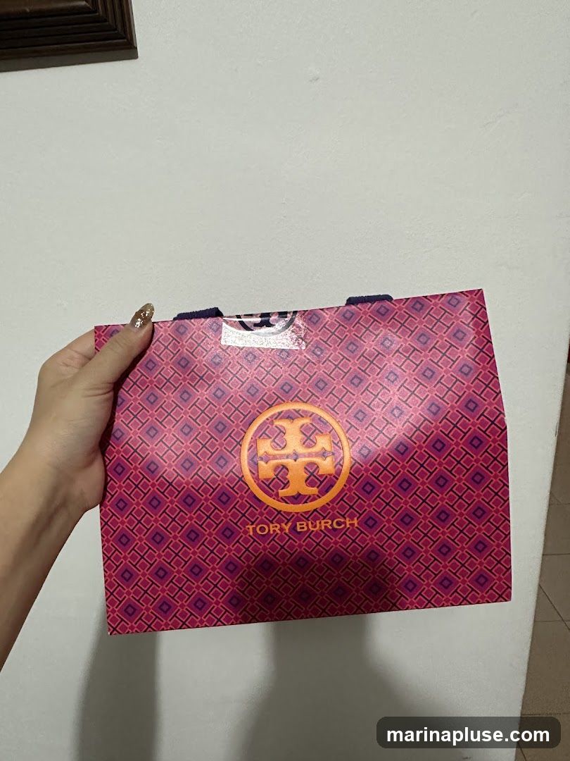 A hand holds a pink and purple patterned Tory Burch shopping bag. The bag features the Tory Burch logo and name in orange.