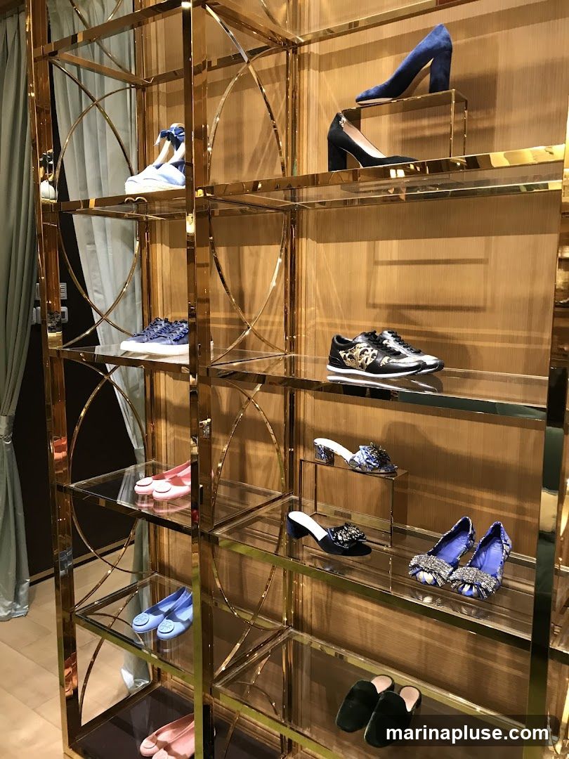 A gold-colored shelving unit displays a variety of women's shoes, including heels, flats, sandals, and sneakers. The shoes are arranged on glass shelves against a wood-paneled wall.