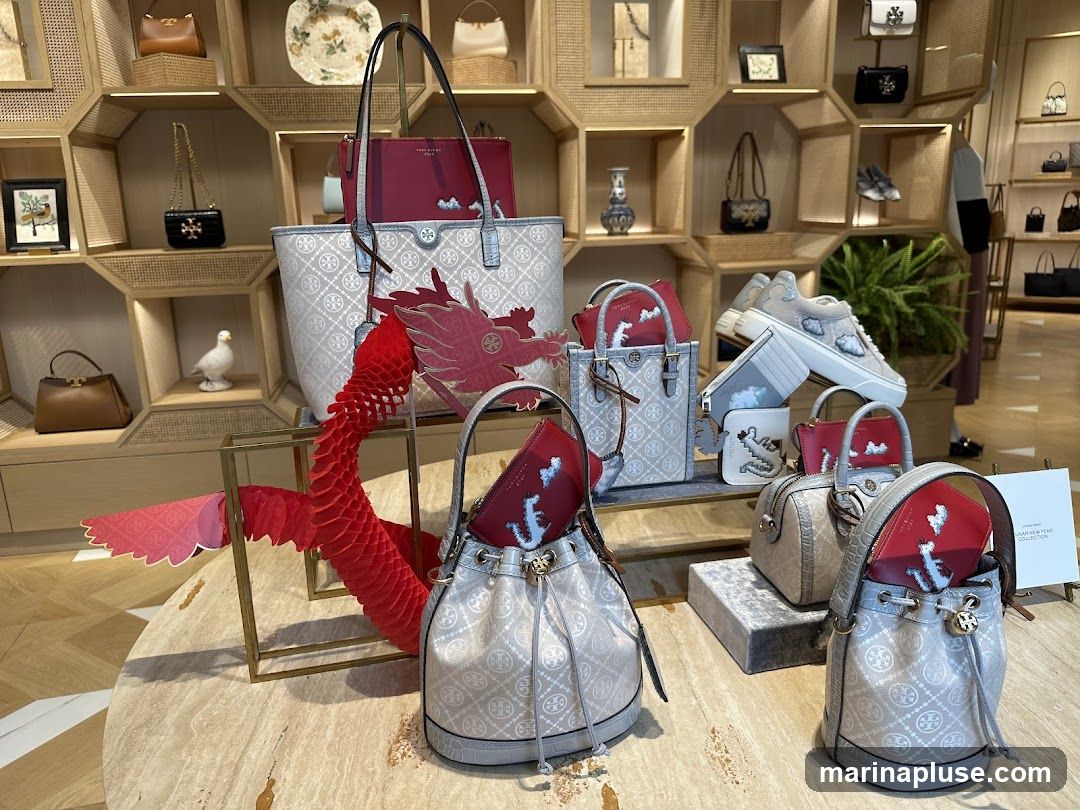 A display of Tory Burch handbags and shoes, featuring a red paper dragon decoration. The collection appears to be themed for the Lunar New Year, with some items featuring cloud and dragon motifs. The bags are in various styles, including totes, bucket bags, and smaller crossbody bags, all in neutral tones with the brand's signature logo pattern. A pair of sneakers with cloud embroidery is also showcased. The display is set against a backdrop of shelving units filled with more Tory Burch merchandise.