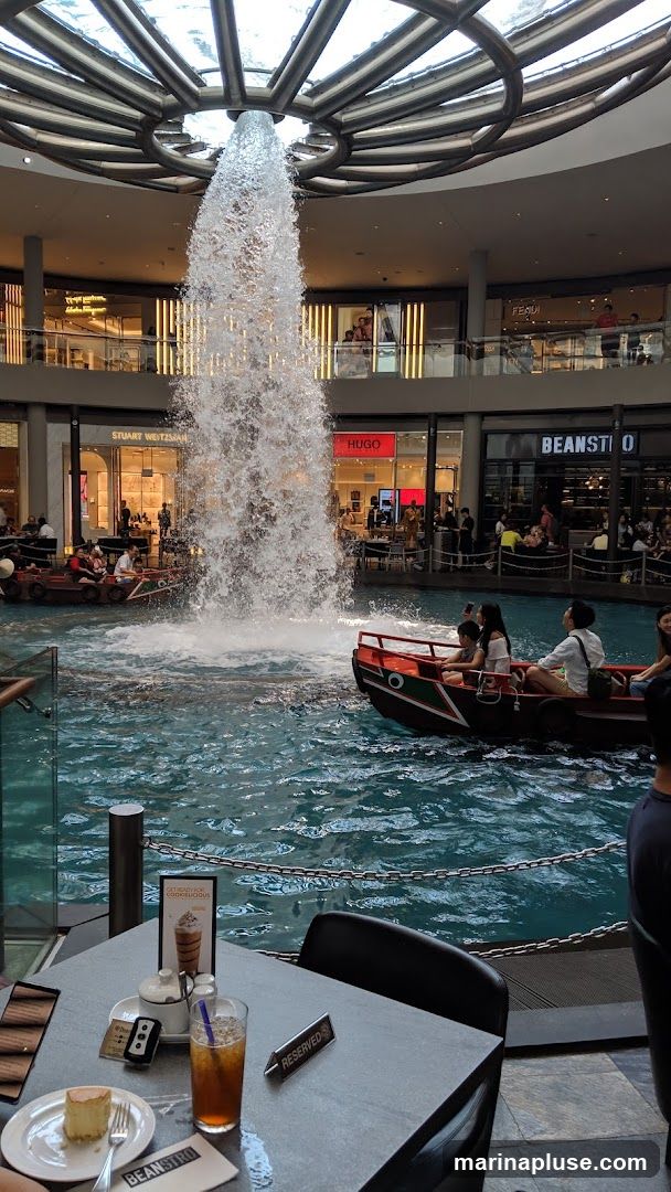 An indoor shopping mall features a large, multi-story waterfall cascading into a pool of water. Small boats with passengers navigate the water. In the foreground, a table is set with a slice of cheesecake, a drink, and a sugar dispenser. Several shops are visible in the background, including Stuart Weitzman, Hugo, and Beanstro.