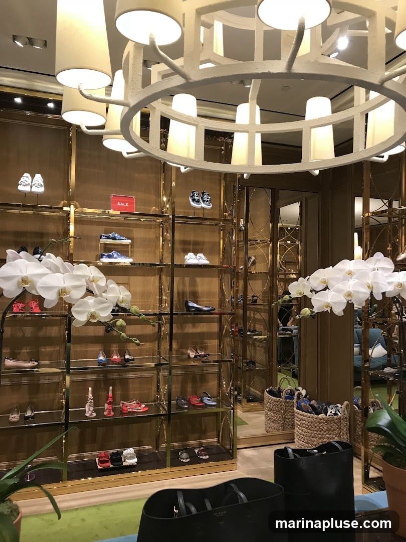 A well-lit interior of a high-end shoe store, featuring gold-accented shelves displaying various women's shoes. White orchids add a touch of elegance. In the foreground, black tote bags are visible. The store appears to be organized and upscale.