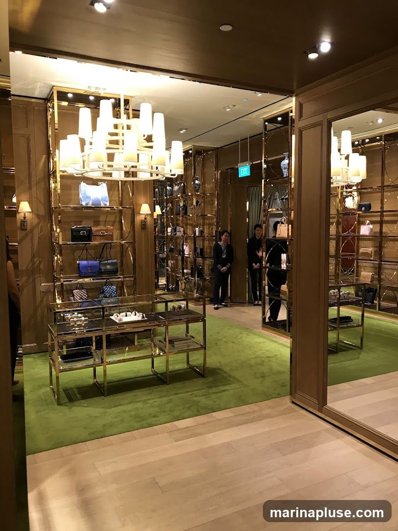 A well-lit, luxurious retail space with gold accents and green carpeting. Shelves display various handbags and accessories. Two staff members are visible in the background. A large chandelier hangs from the ceiling.