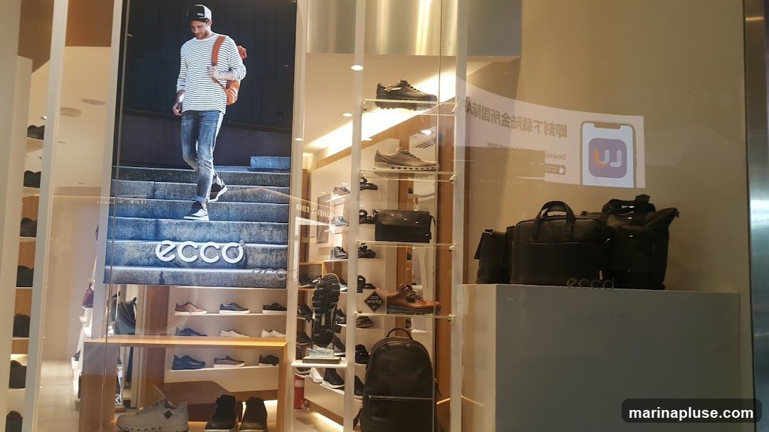A display window for an Ecco shoe store. A large advertisement features a man walking down stairs, with the Ecco logo visible. Inside the window, shelves display various Ecco shoes and bags. A sign with a QR code and app logo is also visible.