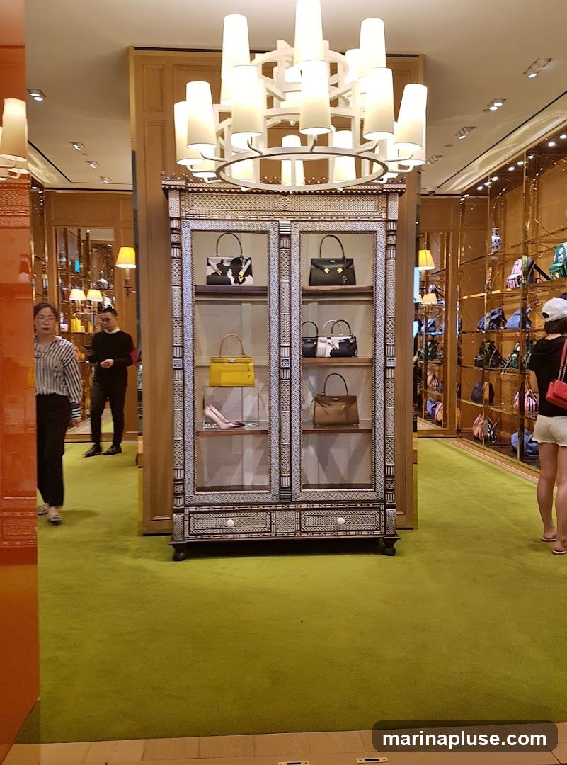 A well-lit interior of a luxury store, featuring a prominent display cabinet filled with various handbags. The cabinet has glass doors and intricate decorative detailing. Shelves inside showcase handbags in different colors and styles, including a bright yellow one and a black one with a distinctive pattern. To the right, more handbags are displayed on gold-colored shelves lining the wall. Two customers are visible in the store, one walking past the display cabinet and another looking at the merchandise on the far wall. A large, modern chandelier hangs from the ceiling, illuminating the space. The floor is covered in a vibrant green carpet.