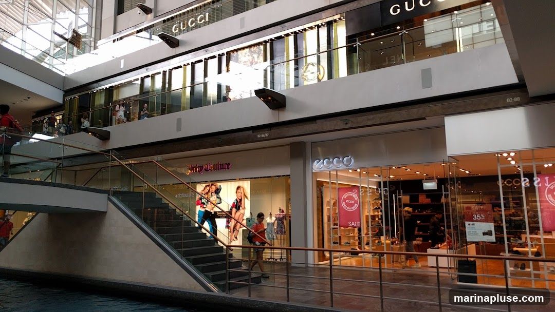 The image shows the interior of a multi-level shopping mall. On the upper level, the Gucci store is visible with its prominent signage. Below, on the lower level, are storefronts for Juicy Couture and Ecco. The Ecco store has a large "SALE 35%" sign displayed in its window. People are seen walking around and browsing the shops. The architecture features modern design with glass railings and escalators.